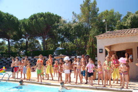 Children and adults in swimwear enjoy a sunny day beside the pool at a glamping site surrounded by trees.