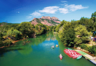 Río con kayakistas y canoas en un alojamiento glamping, rodeado de árboles y montañas bajo cielo azul.