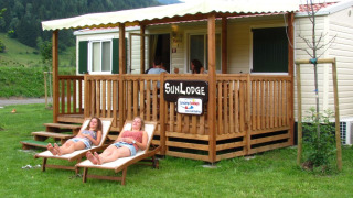 Twee vrouwen relaxen op ligstoelen voor SunLodge Maple, een houten lodge te midden van groen.