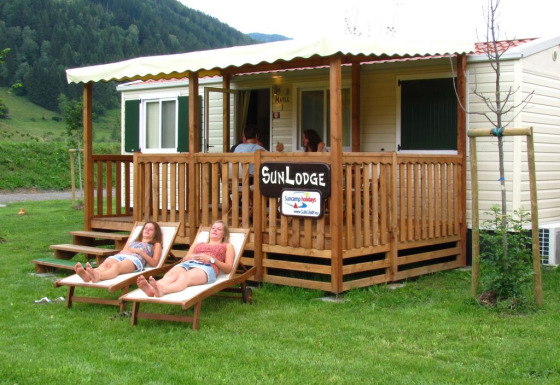 Twee vrouwen relaxen op ligstoelen voor SunLodge Maple, een houten lodge te midden van groen.