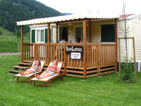 Two women relaxing on loungers outside SunLodge Maple, a wooden porch lodge surrounded by lush greenery.
