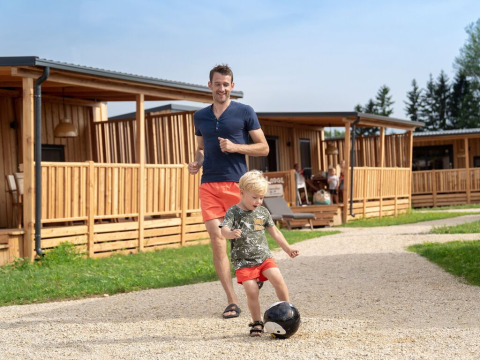 Un homme et un enfant jouent au football devant des lodges en bois à SunLodge BigLeaf sous le soleil.