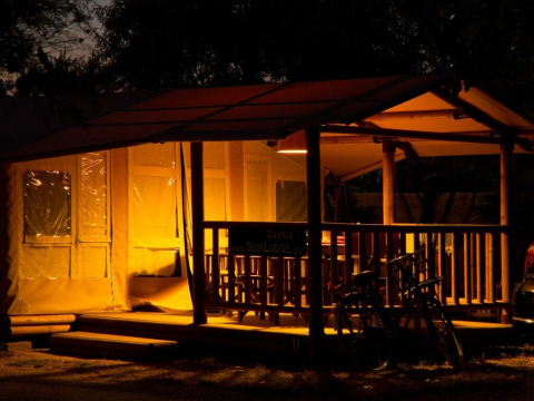 Tienda safari iluminada SunLodge Jungle por la noche con porche y bicicletas en Camping Vilanova Park, España.