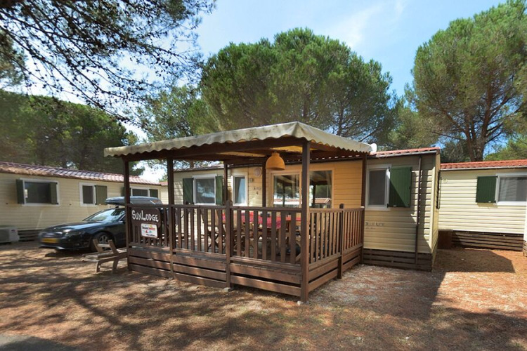 SunLodge Aspen con terraza de madera cubierta y asientos exteriores, rodeada de altos pinos verdes.