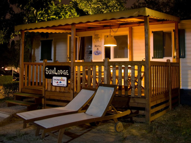 Vista nocturna de SunLodge Maple en Camping Leï Suves, Francia, con terraza iluminada y tumbonas afuera.