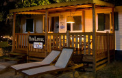Night view of SunLodge Maple at Camping Leï Suves in France, showing a lit porch and two loungers outside.