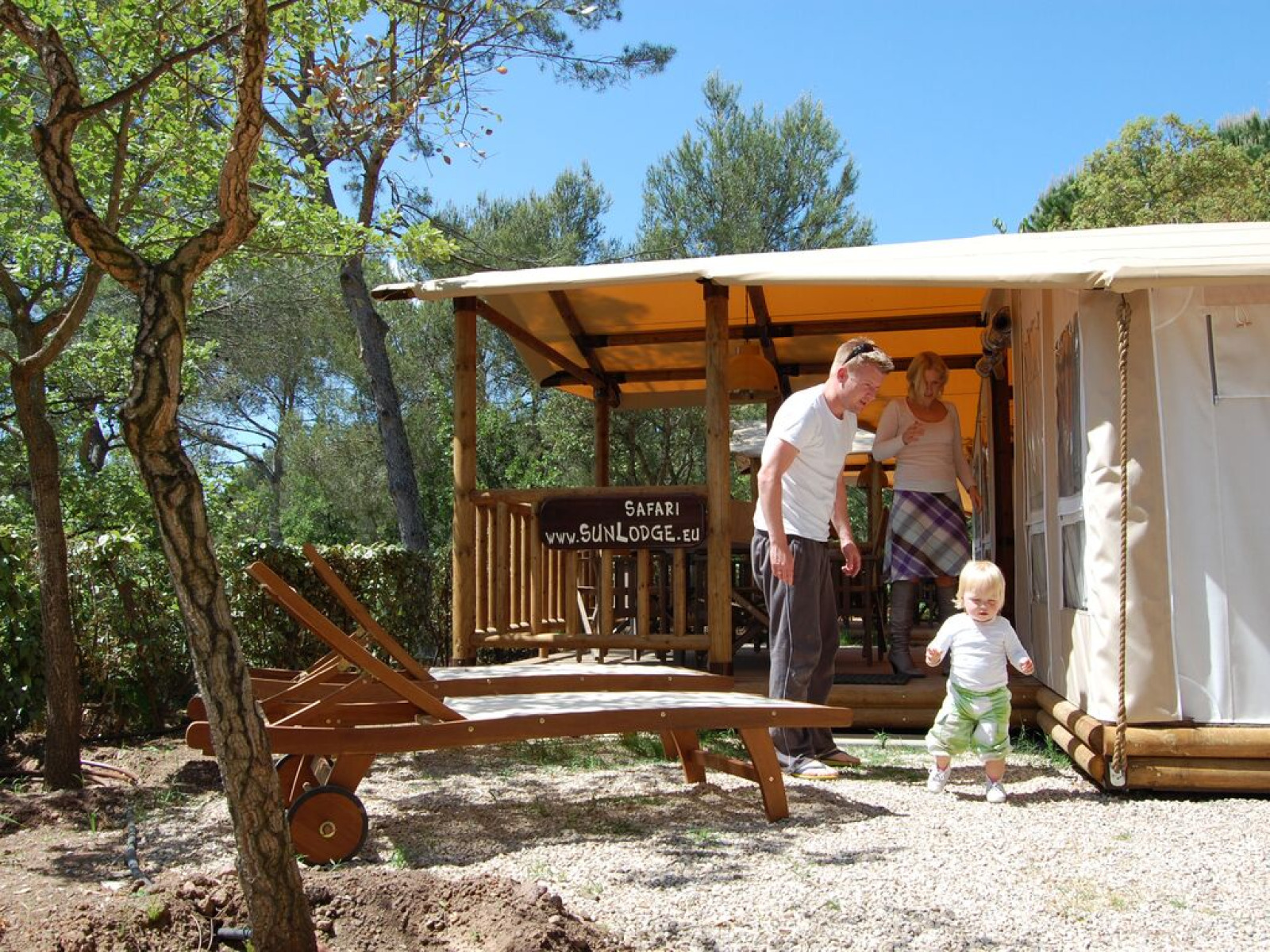 Gezin geniet buiten voor de SunLodge Safari-tent op Camping Leï Suves, omgeven door bomen in Frankrijk.