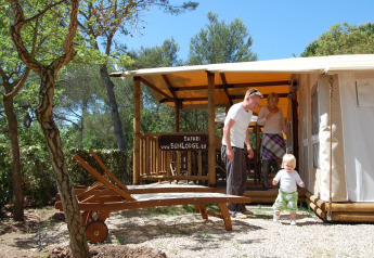 Gezin geniet buiten voor de SunLodge Safari-tent op Camping Leï Suves, omgeven door bomen in Frankrijk.