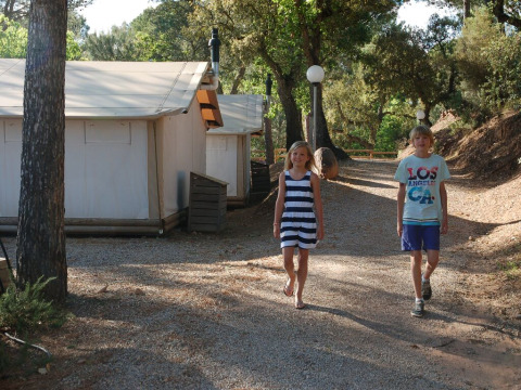 To børn går på stien foran SunLodge Safari-teltet på Camping Leï Suves i Frankrig, omgivet af træer.