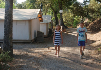 To børn går på stien foran SunLodge Safari-teltet på Camping Leï Suves i Frankrig, omgivet af træer.
