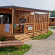 Solrig træhytte kaldet SunLodge BigLeaf med terrasse og liggestol på en grussti omgivet af grønne planter.