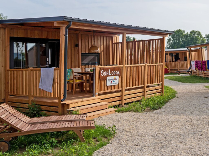 Wooden SunLodge BigLeaf cabin with a shaded porch, lounge chairs, and a gravel path in a tranquil setting.