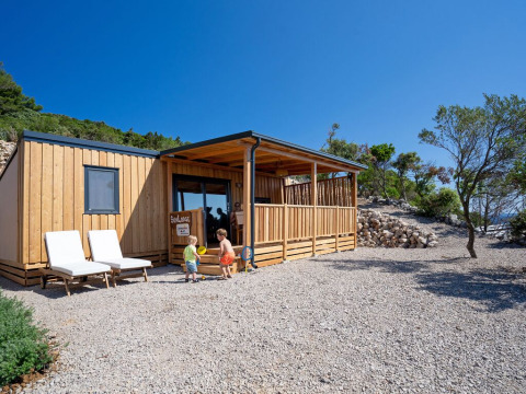 Dos niños juegan frente a SunLodge BigLeaf, una cabaña de madera con porche, rodeada de árboles y piedras.