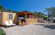 Dos niños juegan frente a SunLodge BigLeaf, una cabaña de madera con porche, rodeada de árboles y piedras.