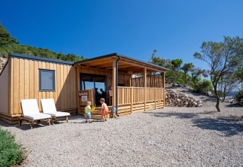 Due bambini giocano davanti a SunLodge BigLeaf, una casetta in legno con veranda tra alberi e rocce.