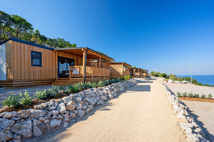 Cabañas de madera modernas alineadas junto a un sendero de grava con vista al mar y cielo despejado.