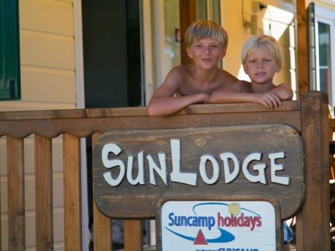 Dos niños sonríen en el porche de SunLodge Maple en Camping Vilanova Park, España, junto a un letrero de madera.
