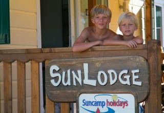 Twee kinderen op het terras van SunLodge Maple in Camping Vilanova Park, Spanje, met een houten bord op de voorgrond.