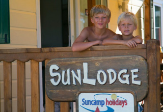 Dos niños sonríen en el porche de SunLodge Maple en Camping Vilanova Park, España, junto a un letrero de madera.