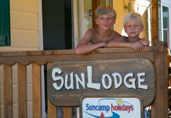 Twee kinderen op het terras van SunLodge Maple in Camping Vilanova Park, Spanje, met een houten bord op de voorgrond.