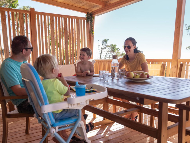 Familia disfruta de una comida en la terraza soleada de madera de SunLodge BigLeaf en Camping Cisano/San Vito, Italia.