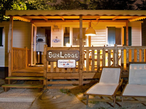 Vista nocturna de SunLodge Redwood en Camping Cisano/San Vito, Italia, con porche iluminado y sillas.