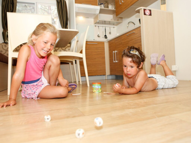 Dos niños juegan con dados en el suelo de una cocina moderna en SunLodge Redwood Premium, Camping Cisano/San Vito, Italia.