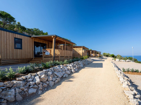 Houten SunLodge BigLeaf lodges langs een grindpad, omgeven door stenen, groen en uitzicht op zee.