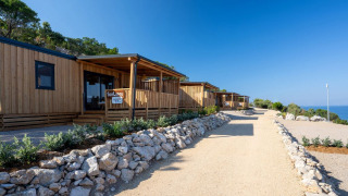 Wooden SunLodge BigLeaf cabins line a gravel path, surrounded by rocks and greenery, overlooking the sea.