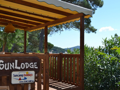 Terraza de madera con cartel 'SunLodge' bajo toldo amarillo, rodeada de árboles verdes y cielo azul.