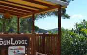 Terraza de madera con cartel 'SunLodge' bajo toldo amarillo, rodeada de árboles verdes y cielo azul.