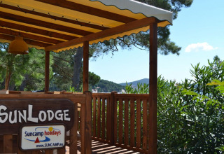Terraza de madera con cartel 'SunLodge' bajo toldo amarillo, rodeada de árboles verdes y cielo azul.