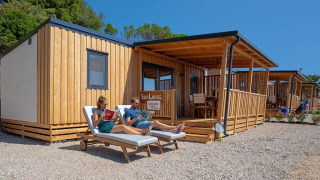 Two people relax on sun loungers outside a SunLodge Catalpa wooden cabin under a clear blue sky.