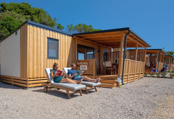 Twee personen ontspannen op ligstoelen voor een SunLodge Catalpa blokhut onder een blauwe lucht.