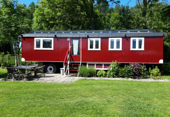 Pipowagen Snuf rojo en Camping De Gouwe Stek, Países Bajos, rodeado de césped, árboles y muebles de exterior.