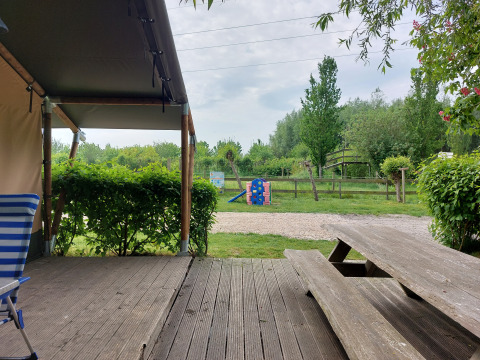 Vista desde la terraza de una tienda safari con banco de madera, parque infantil y vegetación en Camping De Gouwe Stek.