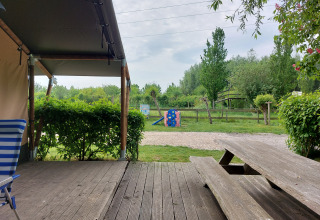 Vista desde la terraza de una tienda safari con banco de madera, parque infantil y vegetación en Camping De Gouwe Stek.
