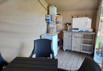 Intérieur d’un lodge avec coin cuisine, table, chaises, réfrigérateur et réchaud sur parquet.