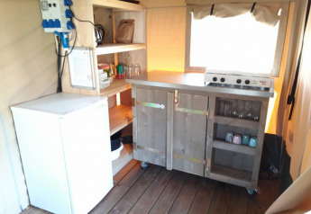 Pequeña cocina con estufa, nevera y estantes abiertos en la Safari tent Merel de Camping De Gouwe Stek.