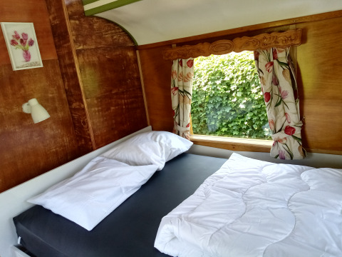 Dormitorio en Glamping Pipowagen Klukkluk con cama doble, cortinas florales y vista natural en los Países Bajos.