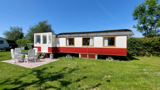 Glamping Pipowagen Klukkluk-Lodge mit Terrasse, weißen Stühlen und Tisch auf einer sonnigen Wiese abgebildet.