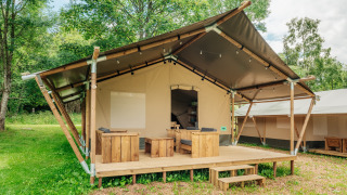 A safari tent with a wooden deck and outdoor seating, surrounded by lush greenery and tall trees.