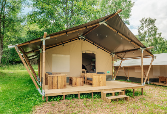 Een safaritent met houten terras en buitenzitjes te midden van groene natuur en bomen.