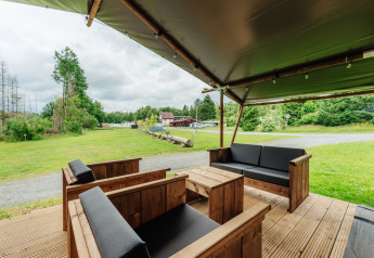 Zona salotto esterna coperta presso una tenda safari con mobili in legno e vista su prato e alberi.