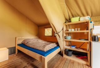 Vista interior de una tienda safari con cama de madera, suelo de madera y estantería abierta con utensilios.