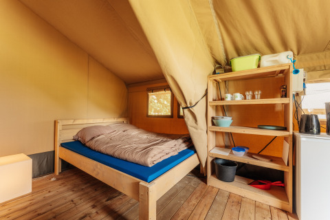 Interior view of a safari tent bedroom with a wooden bed frame, wooden floors, and open shelving unit.