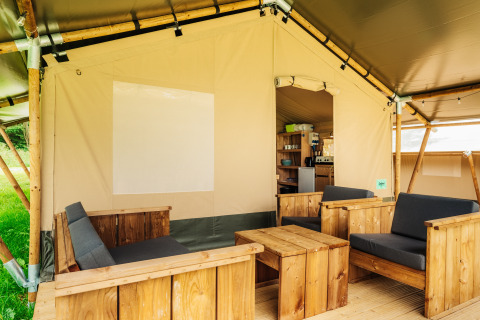 Safari telt loungeområde med træmøbler og bløde hynder, åbent mod et køkken og grøn udsigt udenfor.