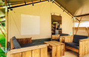 Espace salon sous tente safari avec meubles en bois, coussins et ouverture sur la cuisine et l’extérieur.