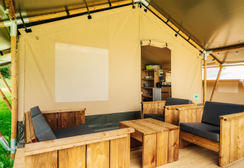 Safari telt loungeområde med træmøbler og bløde hynder, åbent mod et køkken og grøn udsigt udenfor.