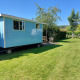 Blå Pipowagen Snuitje-lodge på grøn græsplæne omgivet af træer under klar blå himmel.